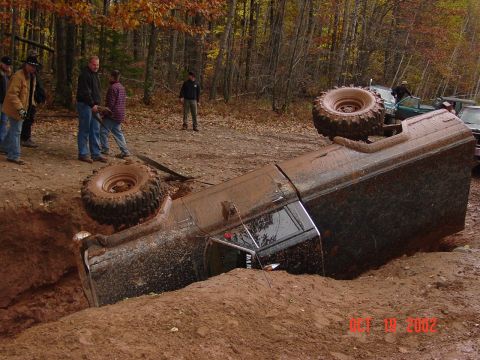 1986 Jeep J-10 - A 1986 Jeep J-10 has rolled onto its side in a muddy off-road pit, with onlookers observing the scene.