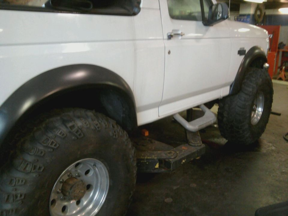 White SUV, possibly a Bronco, with large off-road tires and fender flares. Side view shows running boards and a lifted suspension