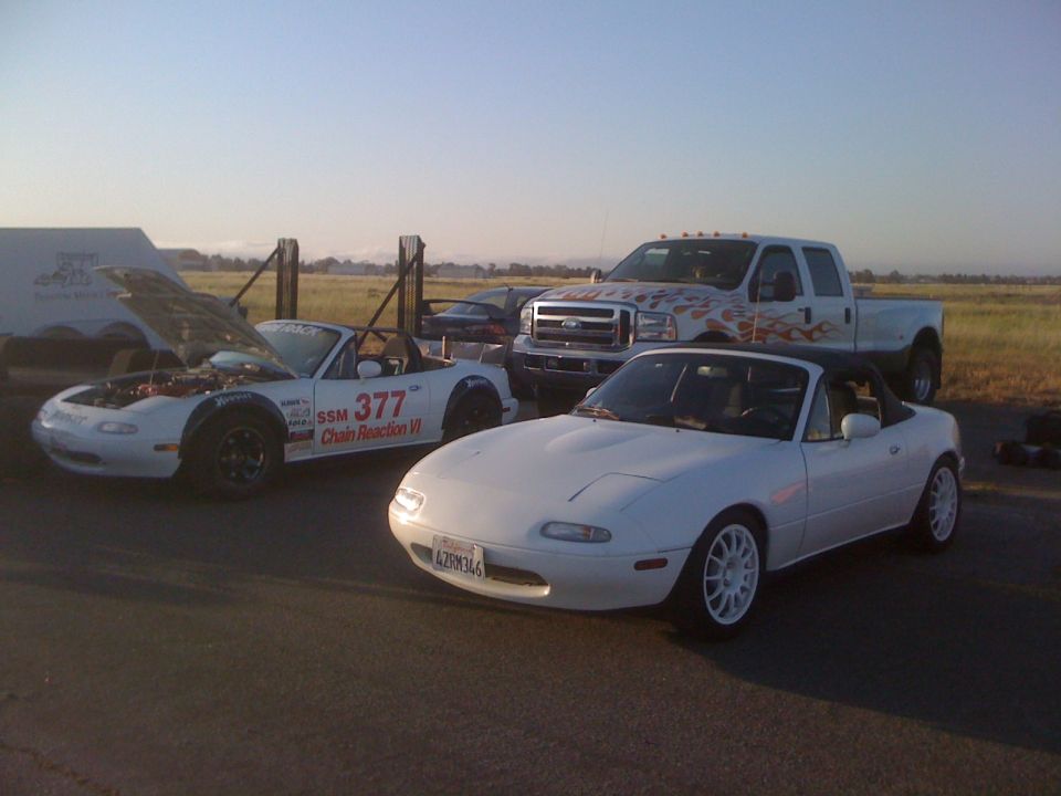 Two white Mazda Miatas, one with hood open, and a white Ford pickup truck with flame decals