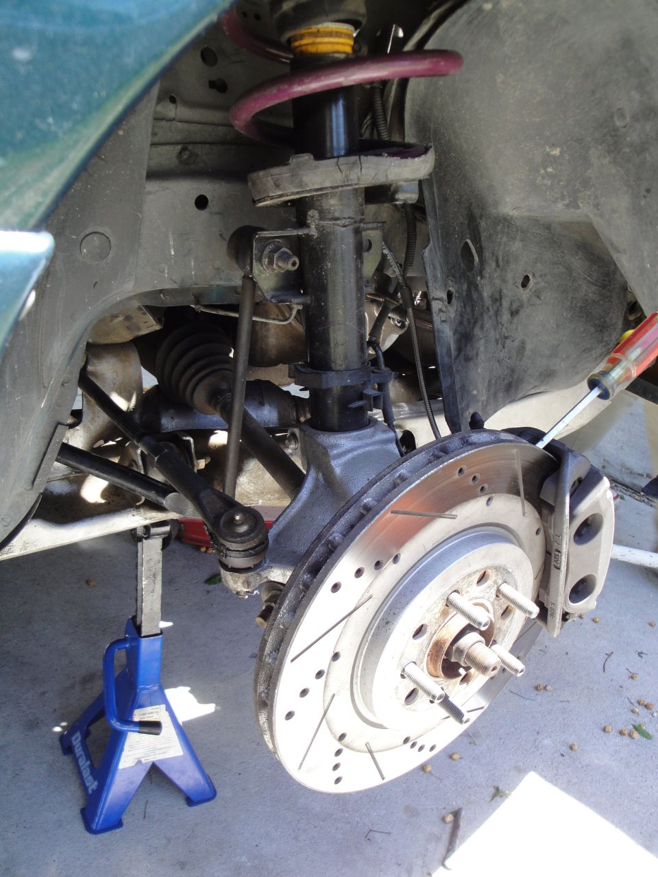Green car's front suspension with a jack stand, perforated brake rotor, and caliper. The car appears to be from the early 2000s