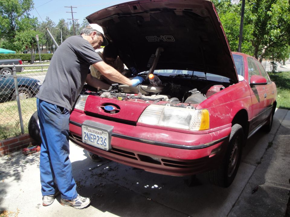 Man working on the engine of a red 1995 Ford Taurus. License plate 2NEP236