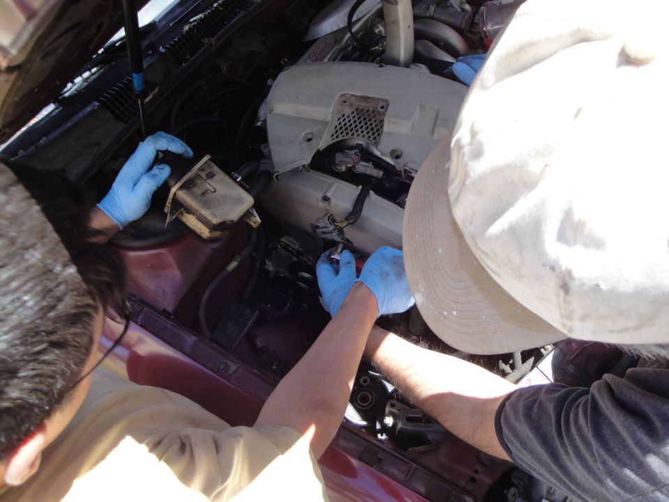 Maroon car engine bay with two people in blue gloves working on engine components