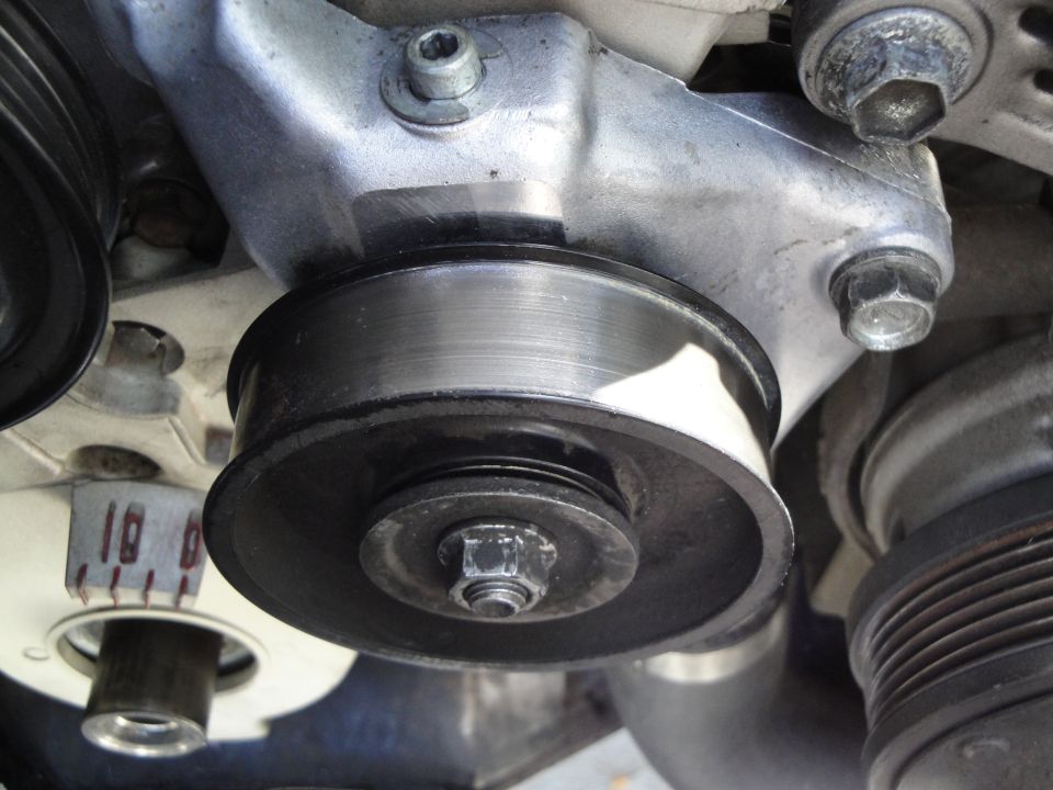 Close-up of an engine pulley with a metal bracket and bolts. A white component with red markings is visible behind it