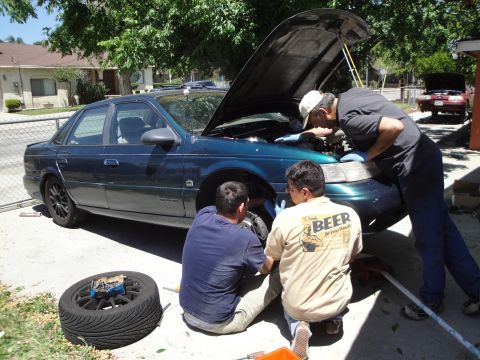 1995 Ford Taurus - Three people are working on a dark green sedan, with the hood up and one wheel removed.
