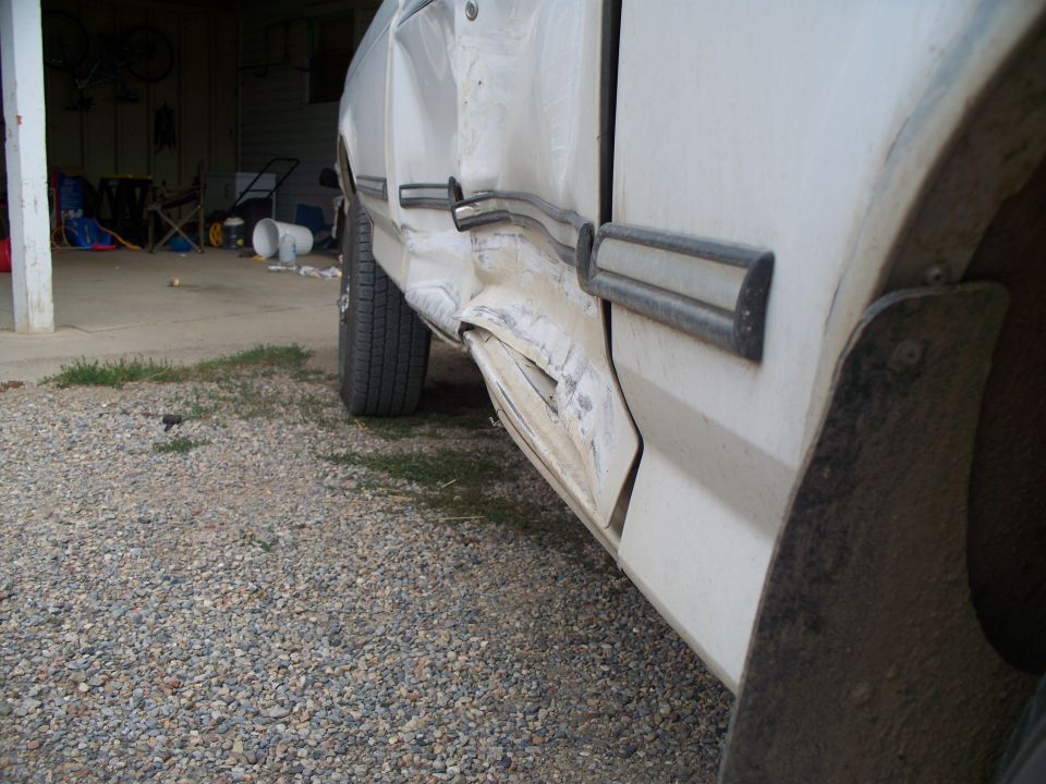 White truck door with significant damage to the lower section and fender, showing rust and body filler