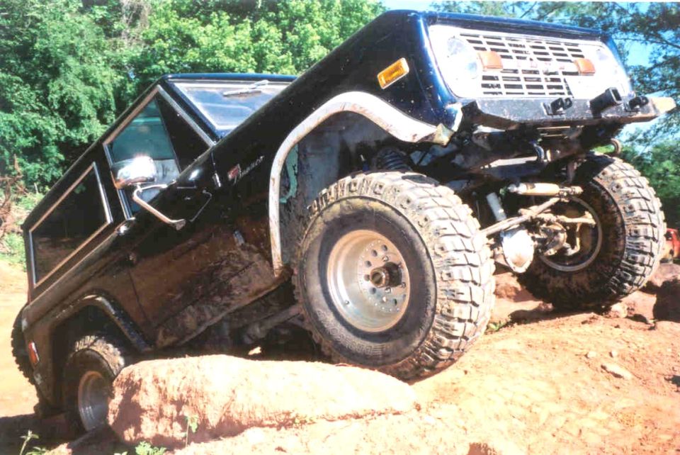 Lifted dark blue Ford Bronco on big mud tires climbing a rock on an off-road trail