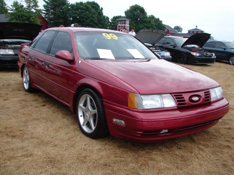 1992 Ford Taurus - A red sedan is parked on a grassy field, with other vehicles visible in the background under a cloudy sky.