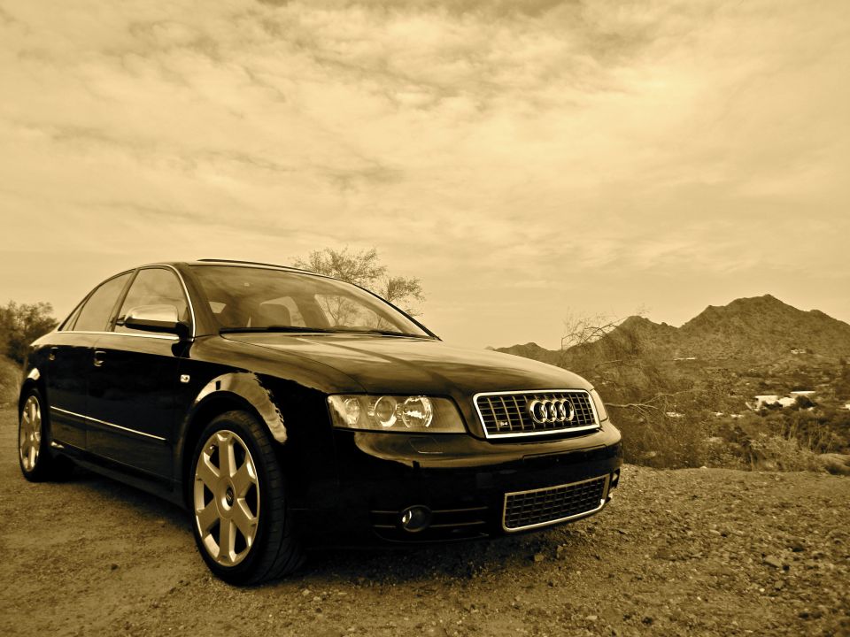 Sepia-toned black Audi sedan photographed low on desert gravel with mountains behind