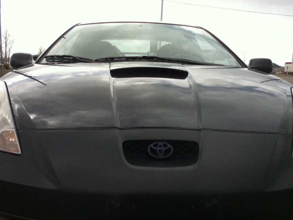Front view of a dark gray Toyota Celica with a hood scoop. The Toyota logo is visible on the grille