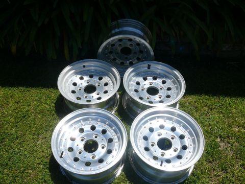 1988 Ford Bronco - Five polished cast-aluminum wheels laid out face-up on a sunlit lawn in front of dark green foliage, each with a round-hole slotted spoke p…