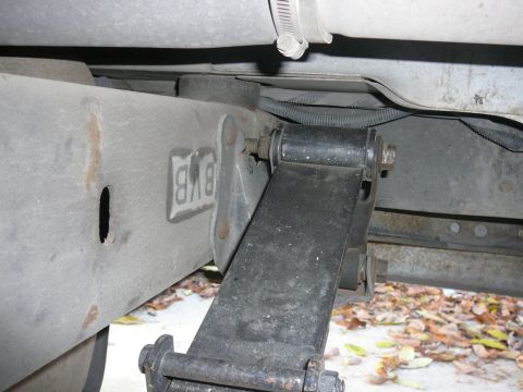 1988 Ford Bronco - Close-up underbody shot of a leaf-spring rear shackle and hanger bracket bolted to a gray boxed frame rail, with the stamped letters 'BVB'…