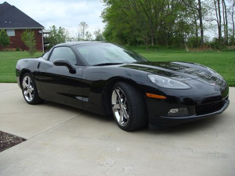 2006 Chevrolet Corvette - A black Chevrolet Corvette is shown from a three-quarter front view, parked on a concrete driveway with a house and trees in the background.