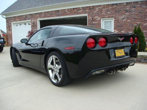 2006 Chevrolet Corvette - Black Chevrolet Corvette with a yellow license plate is parked outside a brick house with a garage.