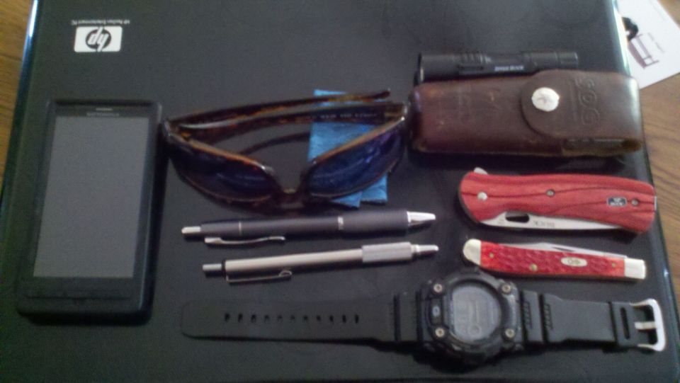 Black smartphone, tortoiseshell sunglasses, two pens, brown leather case with flashlight, red-handled pocket knife, and a digital watch on a dark surface