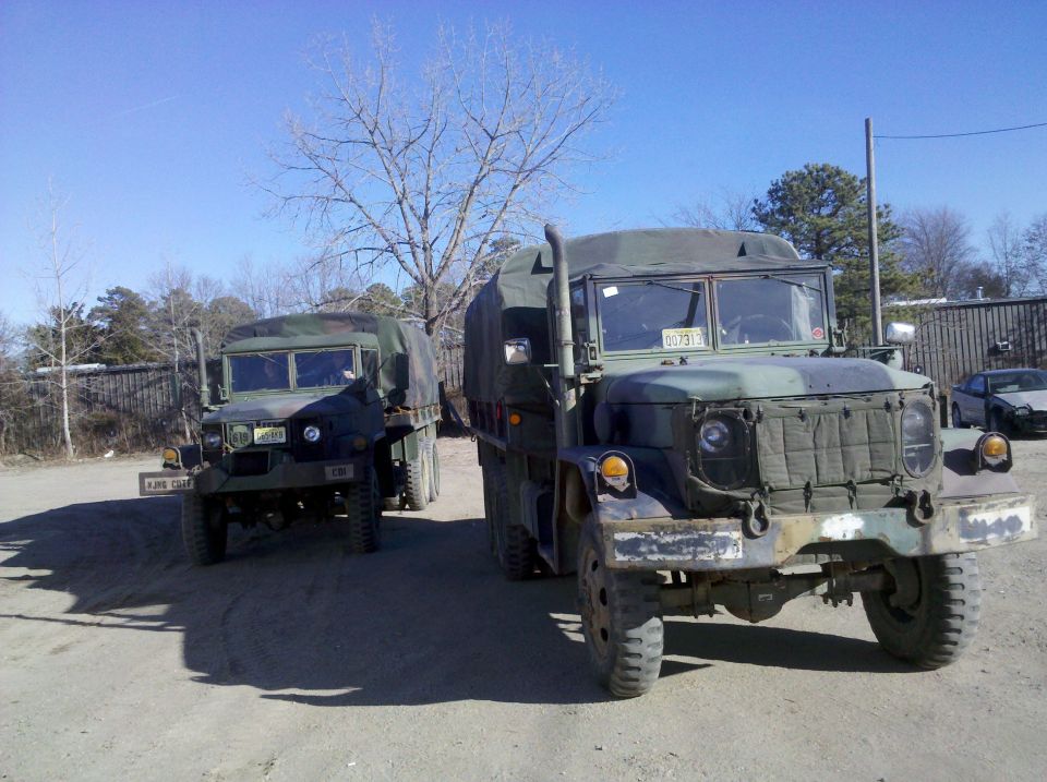 Two military trucks parked outdoors under a clear blue sky. The truck in the foreground is larger with a camouflage tarp cover