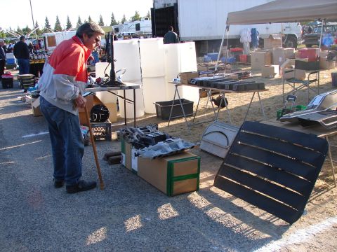1973 Ford Ranchero - Man inspects engine parts at a swap meet with various items for sale in the background.