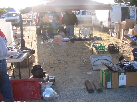 1973 Ford Ranchero - A variety of auto parts are displayed on tables at an outdoor swap meet, with vehicles visible in the background.