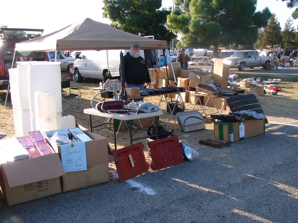 Outdoor swap meet scene with tables of car parts, boxes, a tent, and several vehicles in the background