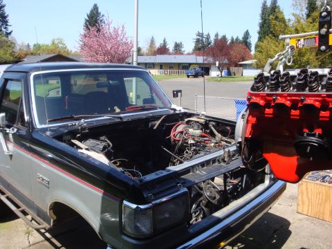 1989 Ford Bronco - A Ford Bronco is undergoing an engine swap, with the old engine removed and a new one suspended by a hoist.