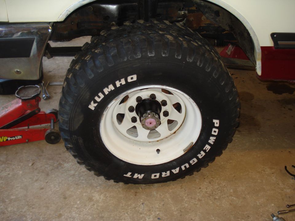 Close-up of a large off-road tire on a white rim, with a red jack visible underneath the vehicle frame