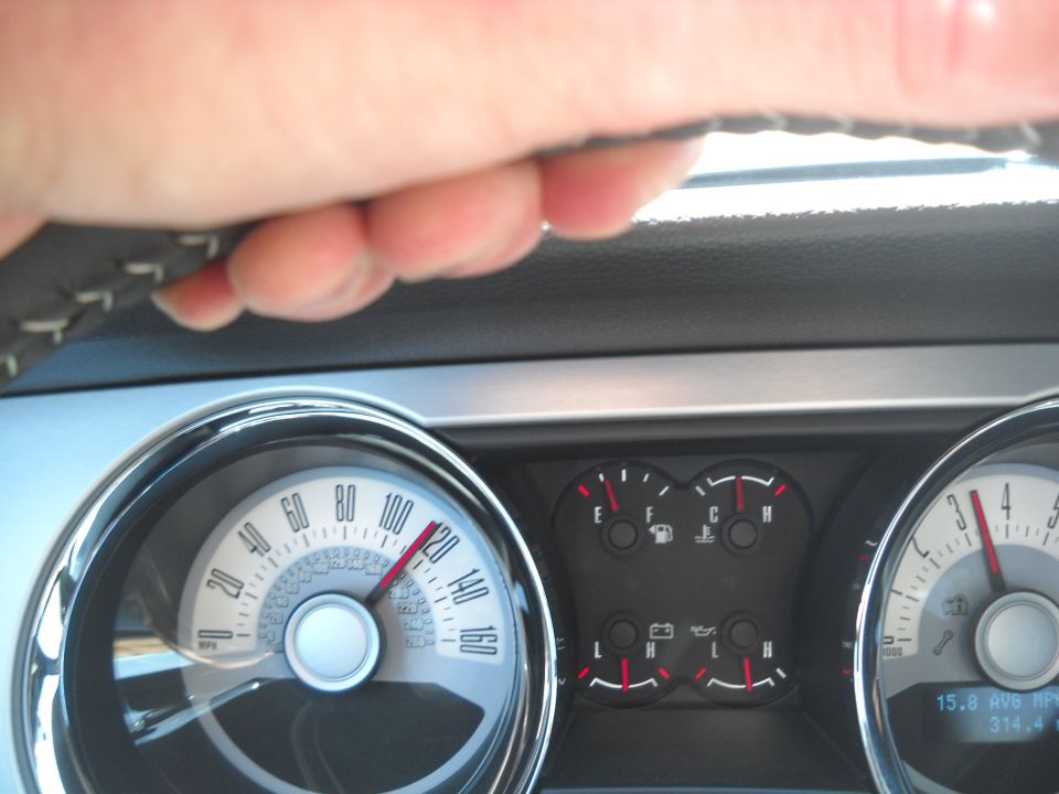 Driver's hand on steering wheel, showing car's speedometer and fuel gauges. Speedometer reads about 115 mph