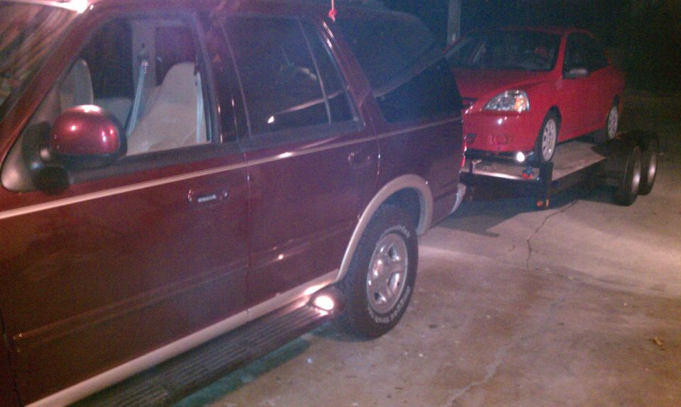 Maroon Ford Expedition side view, towing a red car on a trailer. The Expedition appears to be from the late 1990s