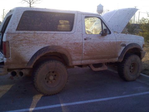 1996 Ford Bronco - This mud-splattered SUV has its hood open, parked on a paved surface with a water tower in the background.