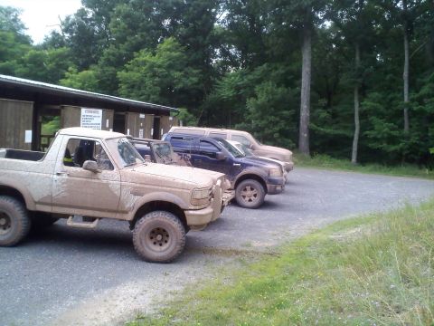 1996 Ford Bronco - Three SUVs, including a mud-splattered 1995 Ford Bronco, are parked in a line on a gravel lot near a wooded area.