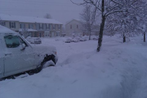 1996 Ford Bronco - A white SUV is partially buried in snow on a street with houses and other vehicles visible in the distance.