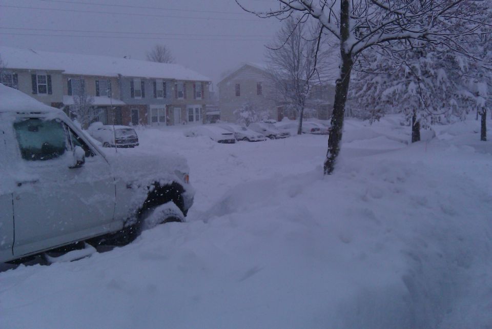 White SUV stuck in deep snow, front quarter view, with houses and other snow-covered vehicles in background