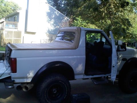 1996 Ford Bronco - White full-size Ford Bronco with a tan soft top, passenger door open and steel wheels, parked among trees in a residential yard.