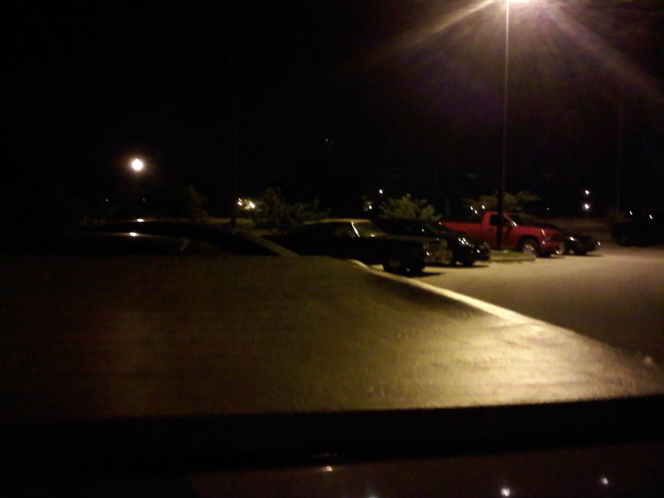 Several cars parked in a lot at night under a streetlamp, with a red truck visible