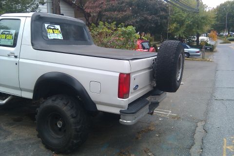 1996 Ford Bronco - Lifted silver full-size Ford Bronco with For Sale signs in the windows, soft top, mud tires and a rear-mounted spare, parked curbside.