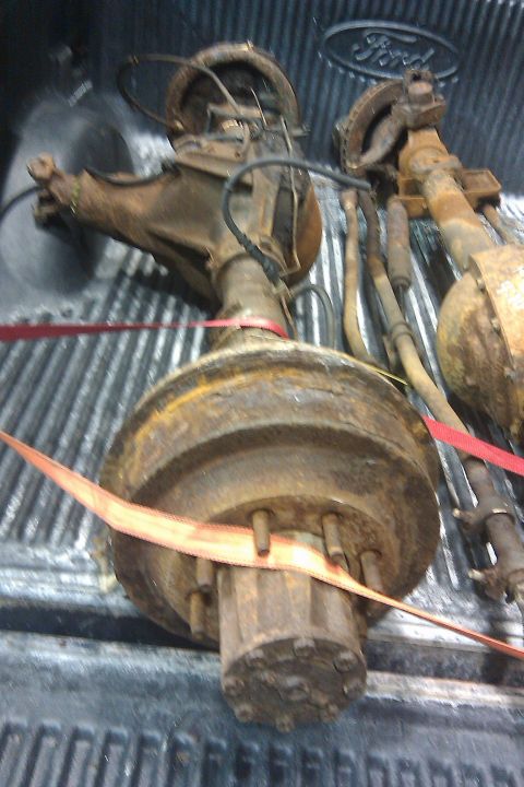 1996 Ford Bronco - Two rusty Ford rear axles are secured with straps in the bed of a truck, with the Ford logo visible in the background.