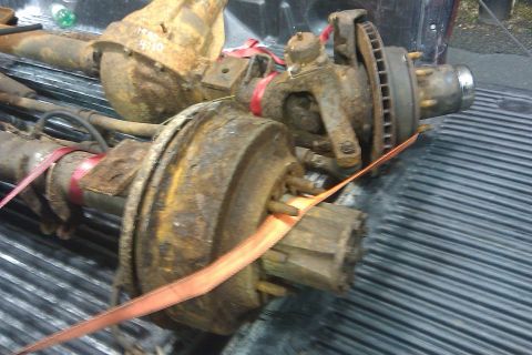 1996 Ford Bronco - Rusty truck axles and suspension parts secured with straps in the bed of a pickup truck.