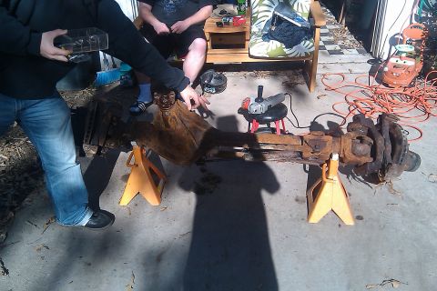 1996 Ford Bronco - A rusty axle assembly is supported by jack stands, with an angle grinder and extension cord nearby, suggesting repair or modification work.