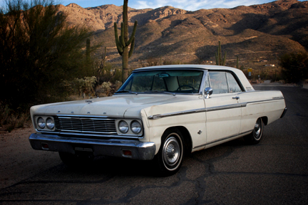 1965 Ford Fairlane - A white 1965 Ford Fairlane coupe is parked in a desert landscape with mountains and saguaro cacti under a blue sky.
