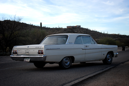 1965 Ford Fairlane - A white 1962 Ford Fairlane coupe is seen from the rear 3/4 angle on a road with a desert landscape.