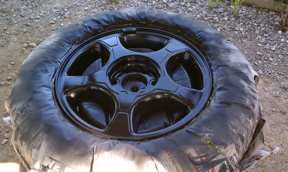 Five-spoke alloy wheel freshly painted gloss black, tire masked with newspaper, lying on gravel