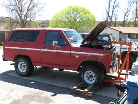 1981 Ford Bronco - Red 1980s Ford Bronco with hood open, being worked on with an engine hoist. Another vehicle is parked behind it.