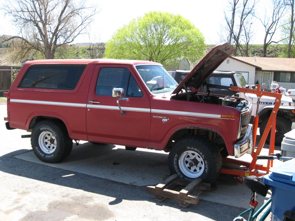 Red 1980s Ford Bronco with hood open, engine bay visible. Orange engine hoist attached. Another white vehicle in background