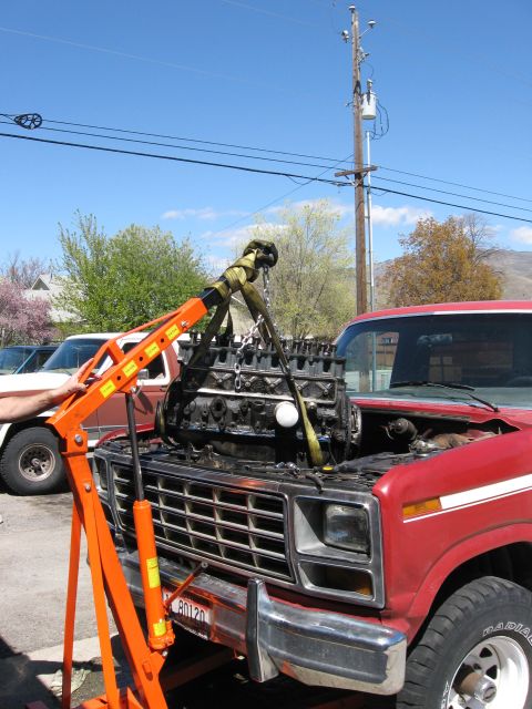 1981 Ford Bronco - An engine is hoisted out of a red Ford truck, revealing its engine bay and front grille.