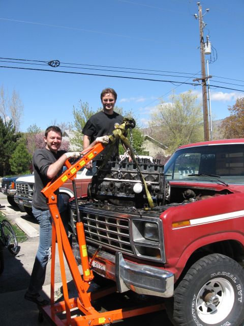 1981 Ford Bronco - Two men are smiling while using an engine hoist to lift an engine into a red 1980s truck, with another truck visible in the background.