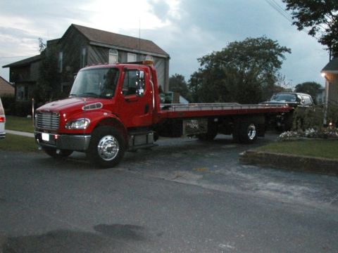 1994 Ford Bronco - A red tow truck is parked on a street, carrying a dark SUV on its flatbed.