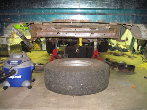 1979 Ford Bronco - The rear of a green vehicle is raised on jack stands, with a spare tire and shop vac nearby.