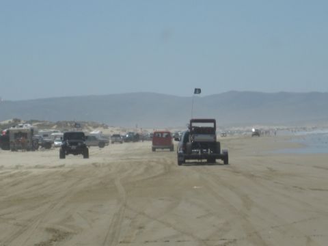 1989 Ford Bronco - Multiple off-road vehicles are seen driving on a sandy beach, with dunes and the ocean in the distance.