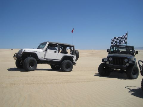 1989 Ford Bronco - Two Jeep Wranglers are parked on sand dunes, one white and one black, under a bright blue sky.