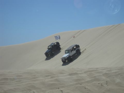 1989 Ford Bronco - Two Jeeps ascend a sandy slope under a clear blue sky, kicking up dust as they climb.