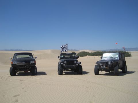 1989 Ford Bronco - Three off-road vehicles are parked in the sand dunes on a clear day.