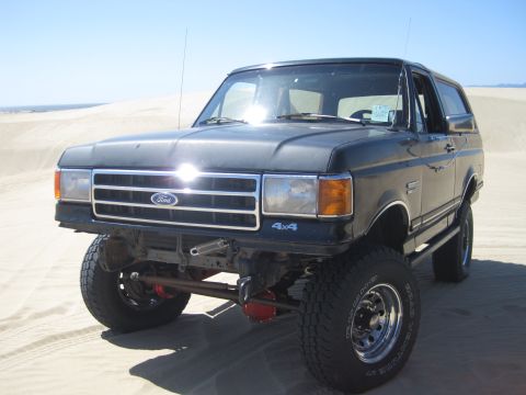 1989 Ford Bronco - A dark gray Ford Bronco 4x4 is parked on sand dunes under a clear blue sky.